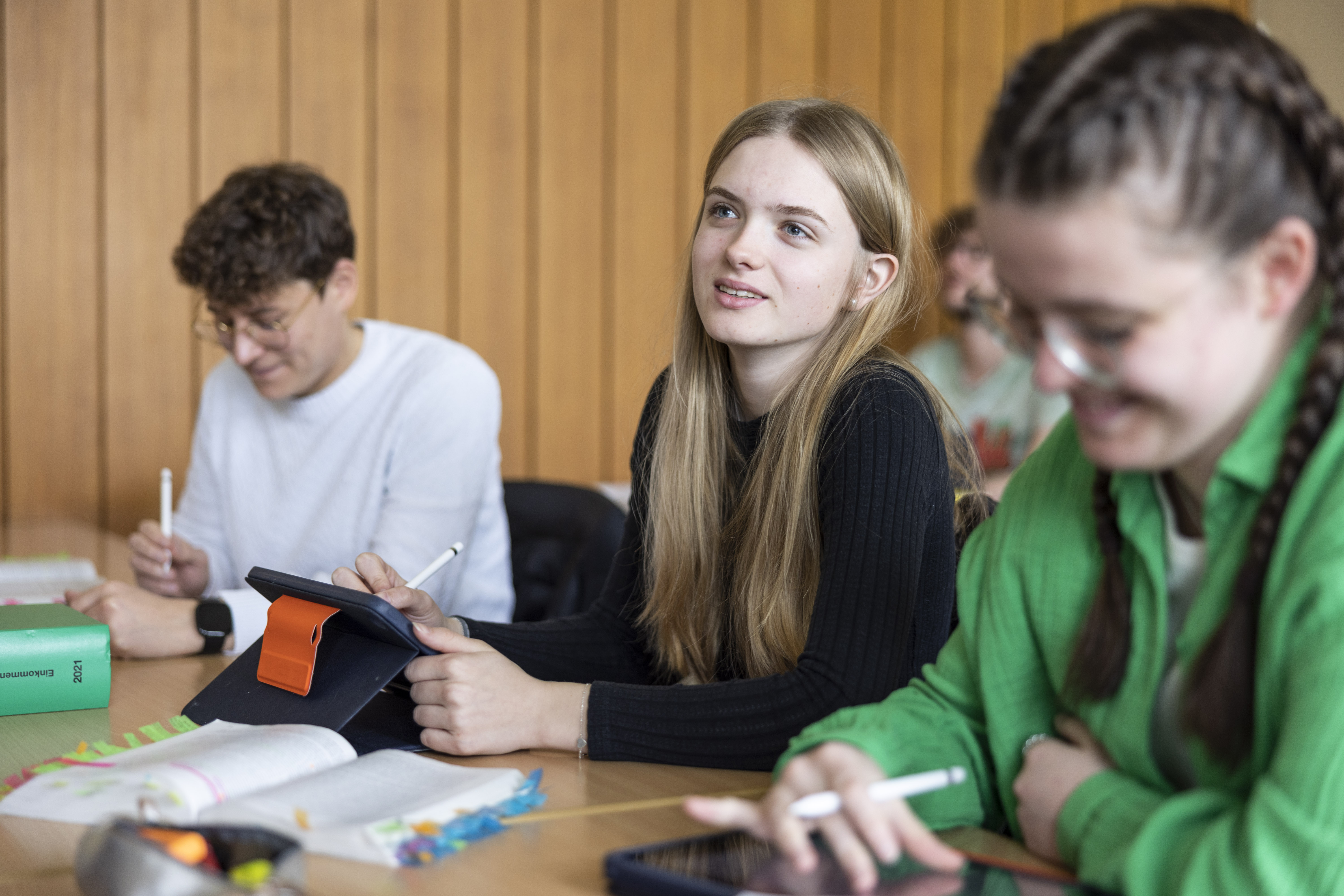 Schülerin sitzt an mit ihrem Tablet in einem Klassenraum und schaut nach vorn.