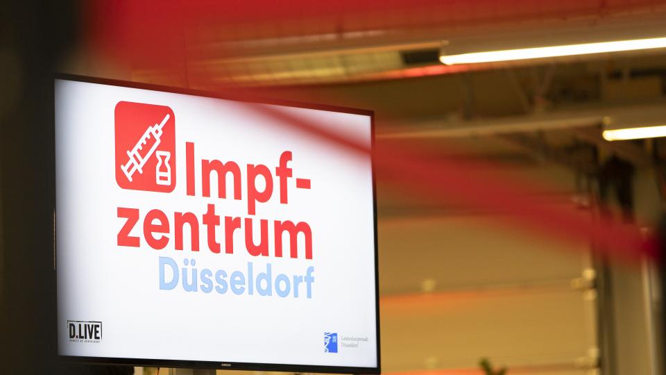 Ein großer Bildschirm in einem Raum, darauf steht "Impfzentrum Düsseldorf".